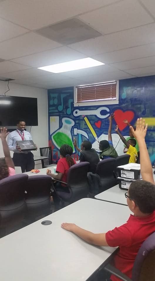 Milan Balinton handing a laptop to students in a classroom, many hands raised