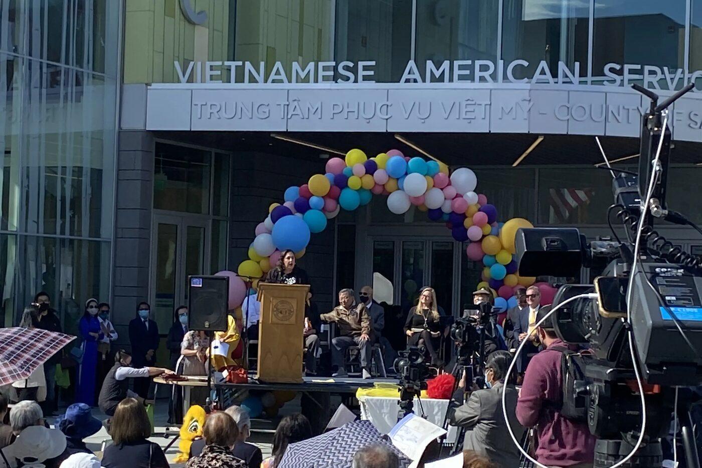 A seated crowd outside the Vietnamese American Service Center in San Jose for a county event, with news cameras covering a speaker at the podium